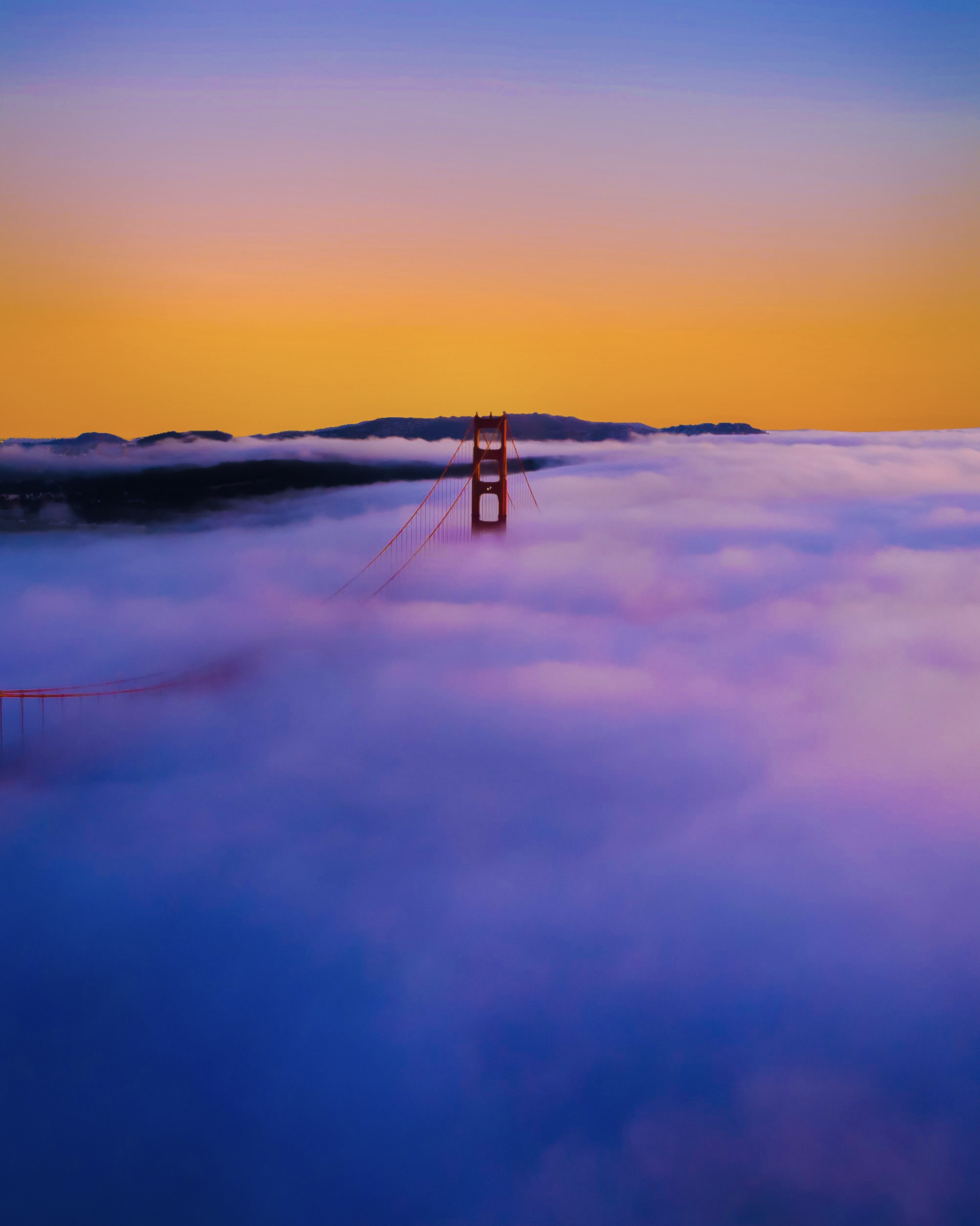 The top of the north tower of the Golden Gate Bridge, peeking out from a sunset-painted fog.